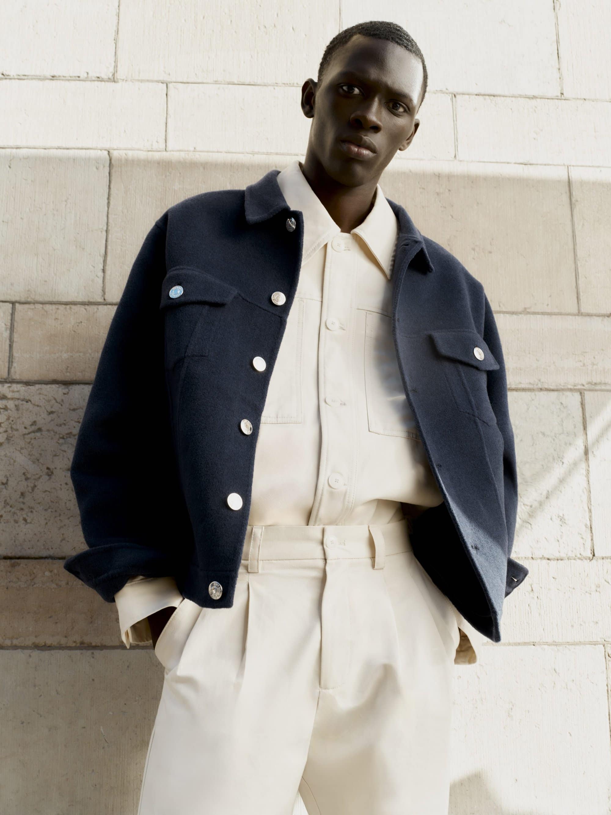 Black man wearing a dark navy wool jacket over an off-white button shirt and trousers against a stone exterior wall.