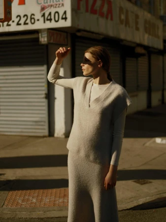 Woman in a knit vest and skirt standing outdoors on a street, shielding her eyes from bright sunlight