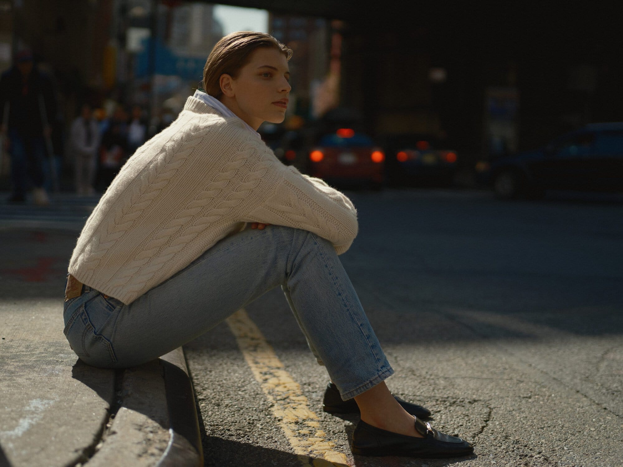 Woman wearing cream cable-knit sweater and light blue jeans sitting on a New York City curb.