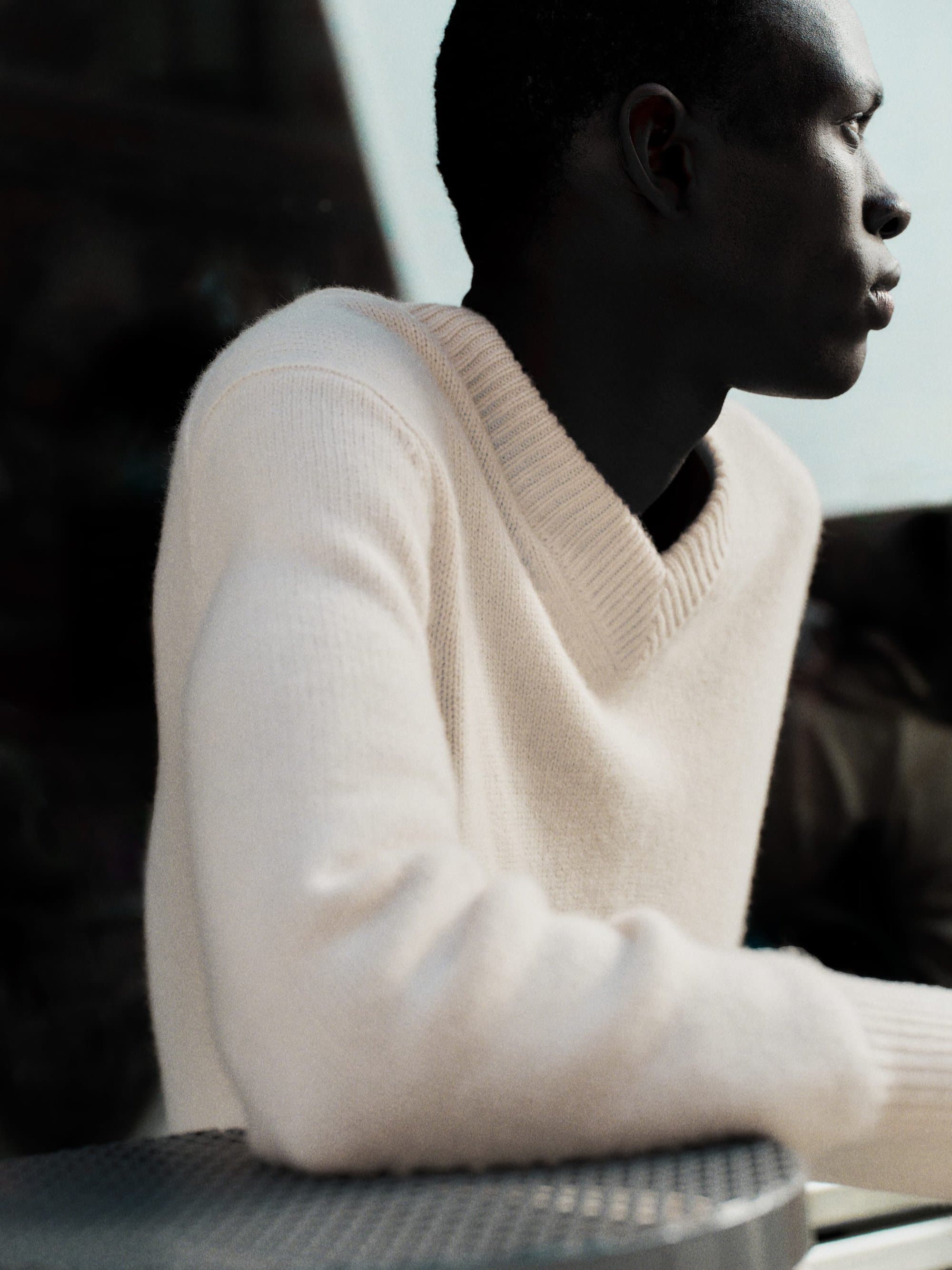 Close-up portrait of a Black man wearing a cream V-neck knit sweater, looking away in profile against a dark background.