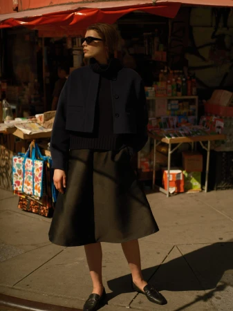 Woman in dark sunglasses wearing a navy cropped jacket, black turtleneck, midi skirt, and loafers on an urban street.