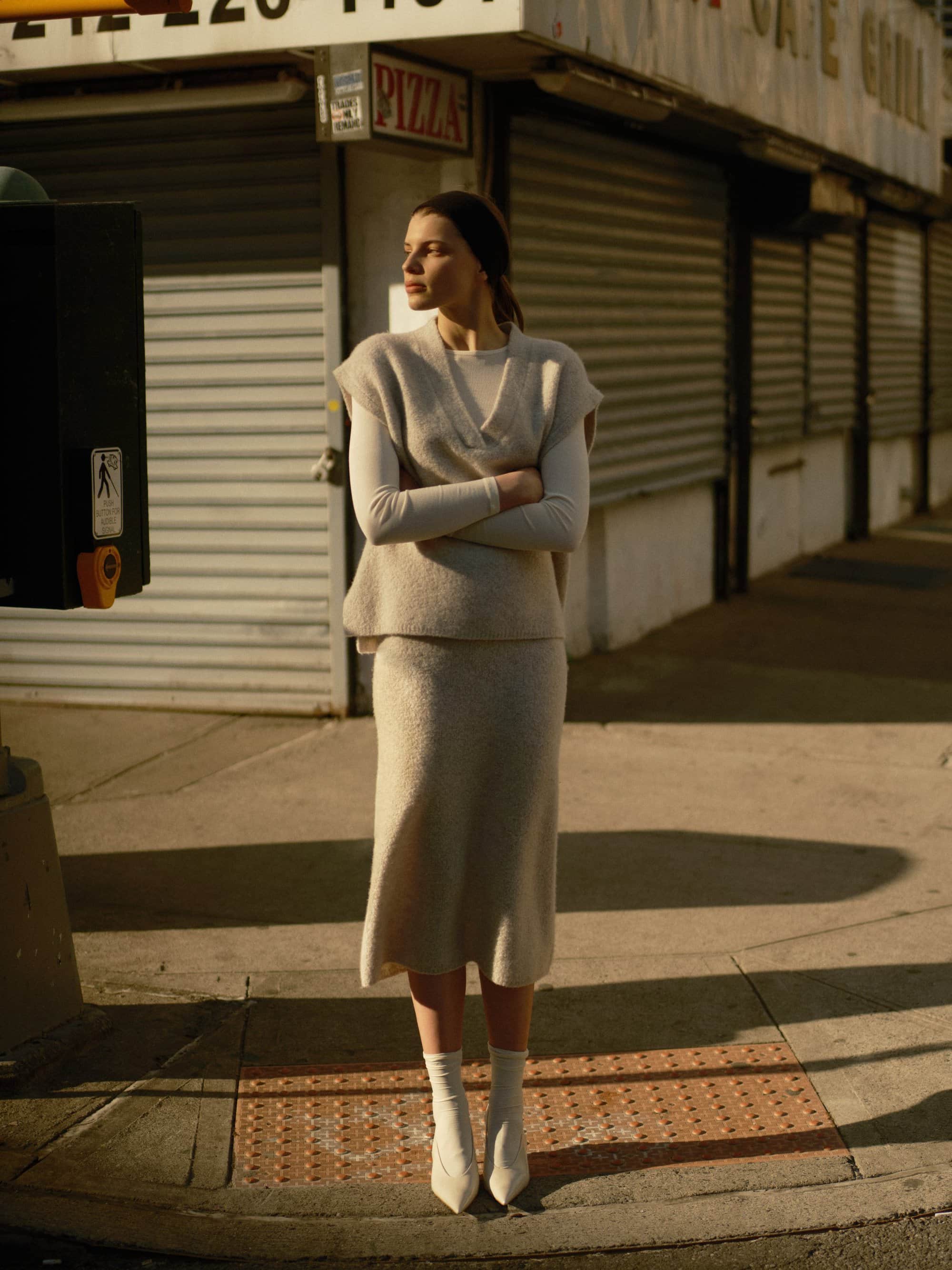 Woman wearing a beige knit vest and skirt standing on a New York City sidewalk in warm afternoon sunlight.