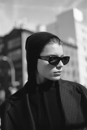 Model wearing dark sunglasses and a black hooded top against an urban background for Soft Goat.