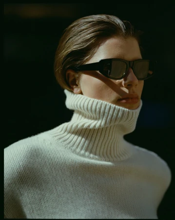Model wearing a white turtleneck sweater and black sunglasses outdoors in natural sunlight