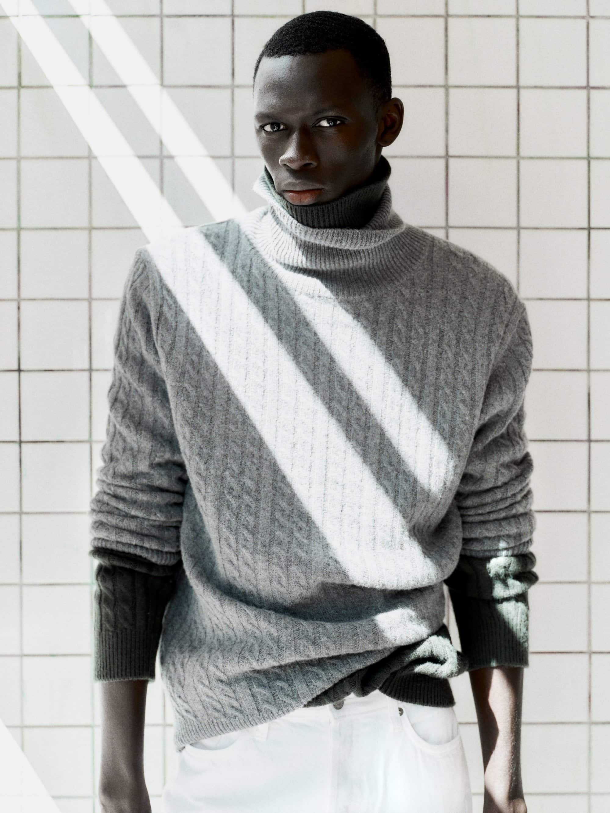 Black man wearing a grey cable knit turtleneck sweater against a white tiled wall with diagonal light streaks