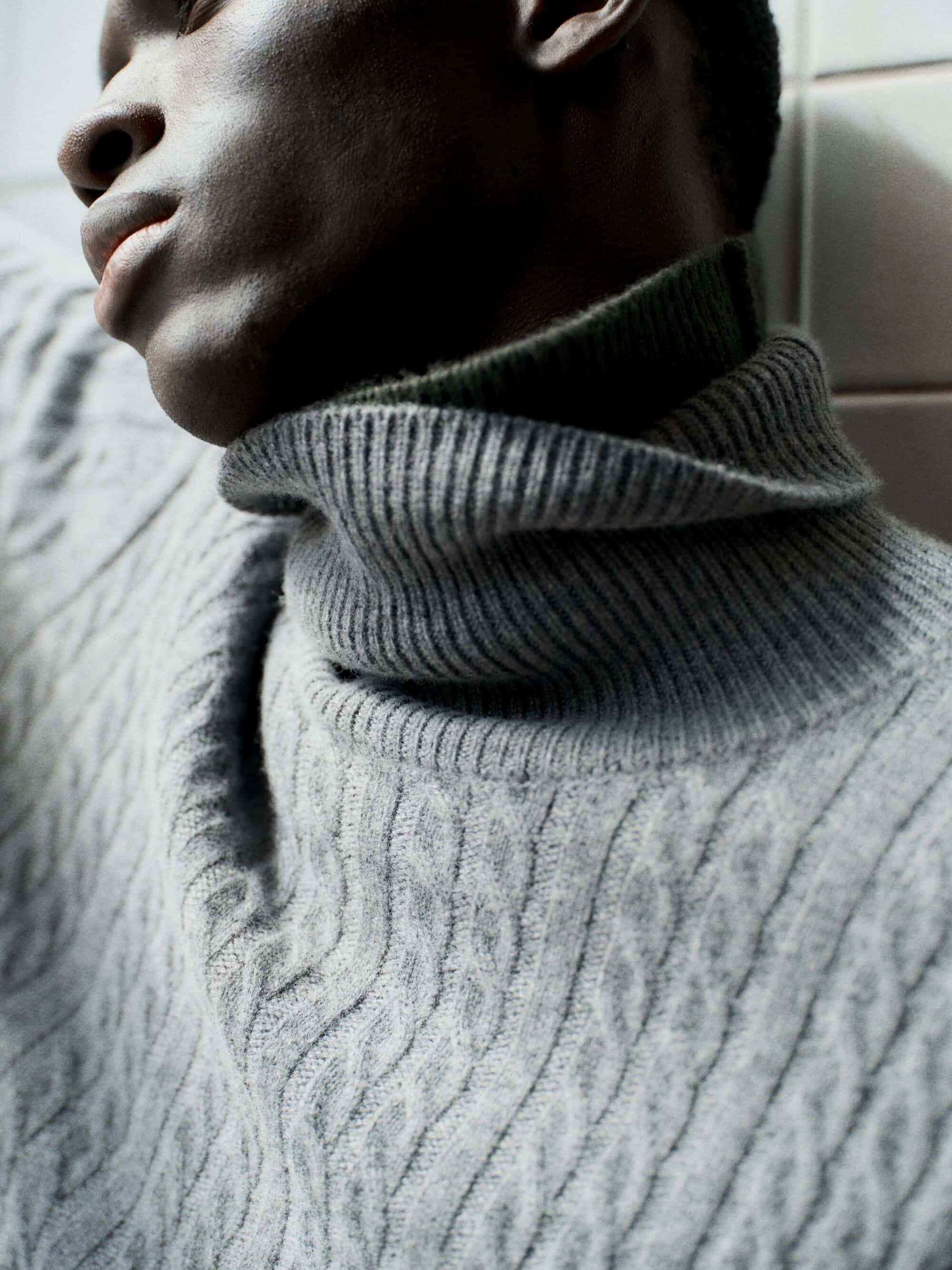 Close-up portrait of a Black man wearing a grey cable knit turtleneck sweater against a tiled wall