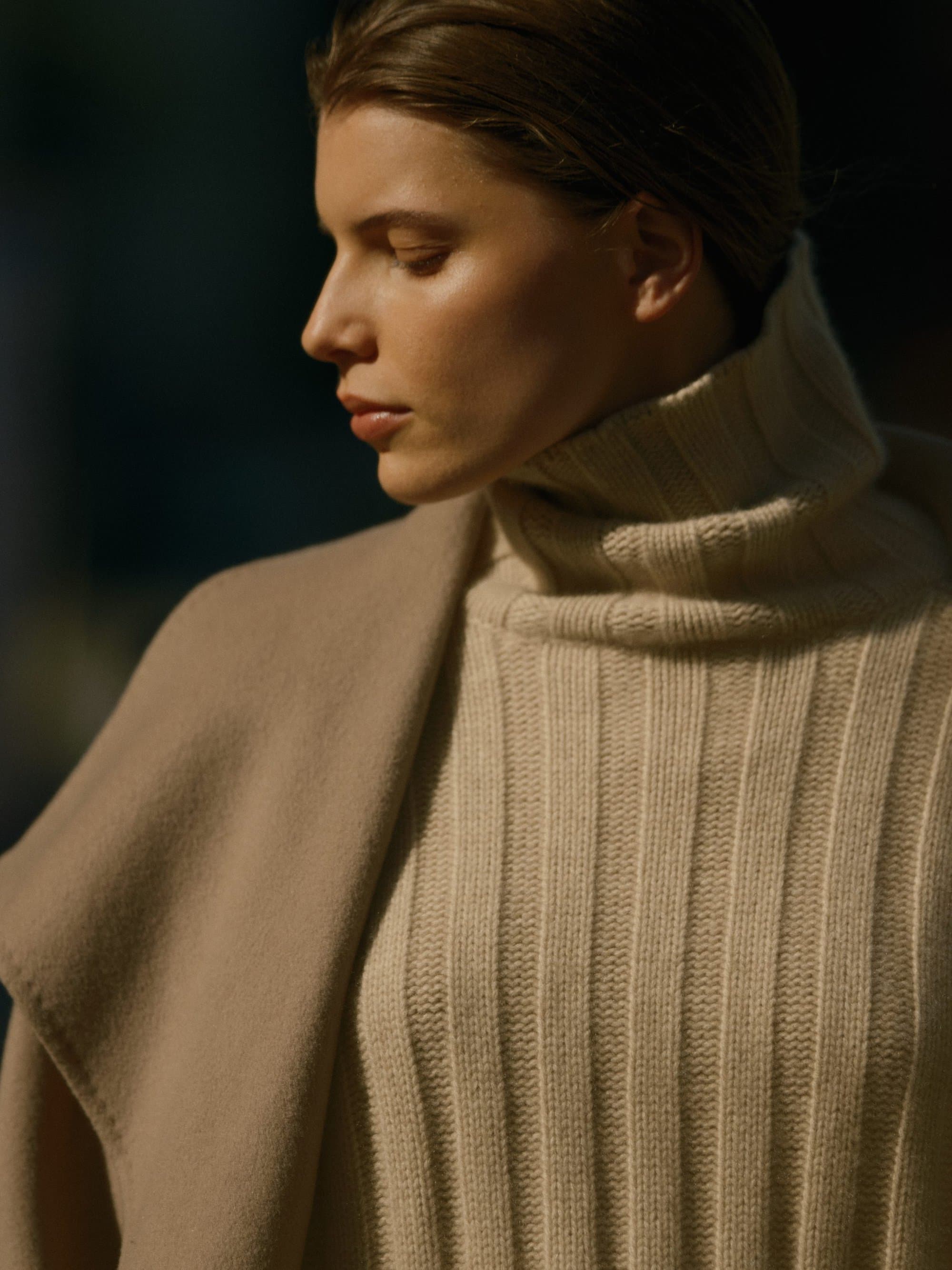 Close-up portrait of a woman wearing a cream ribbed turtleneck sweater and draped camel coat in warm daylight