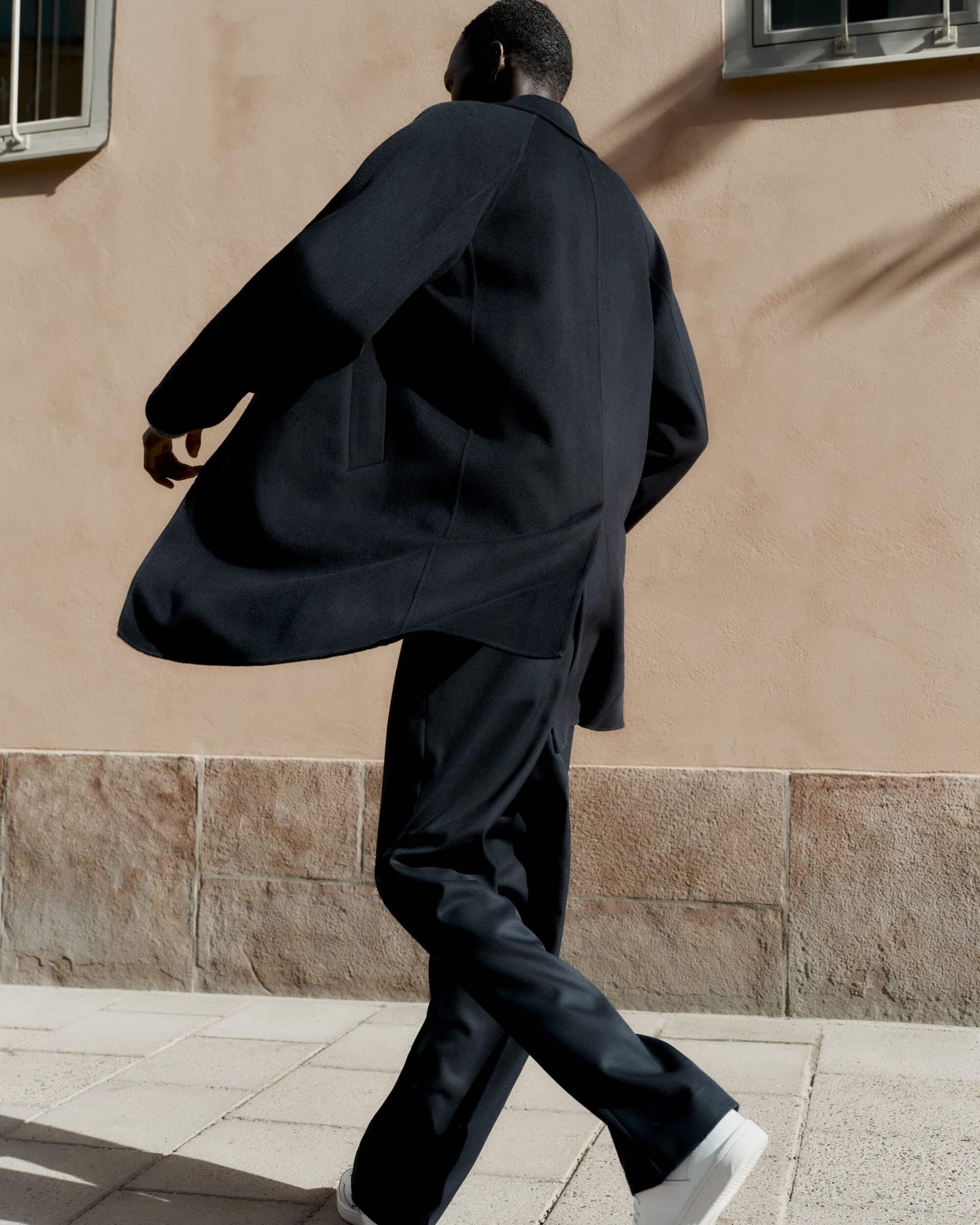Man wearing a dark coat and trousers walking past a peach-colored building exterior in New York City.
