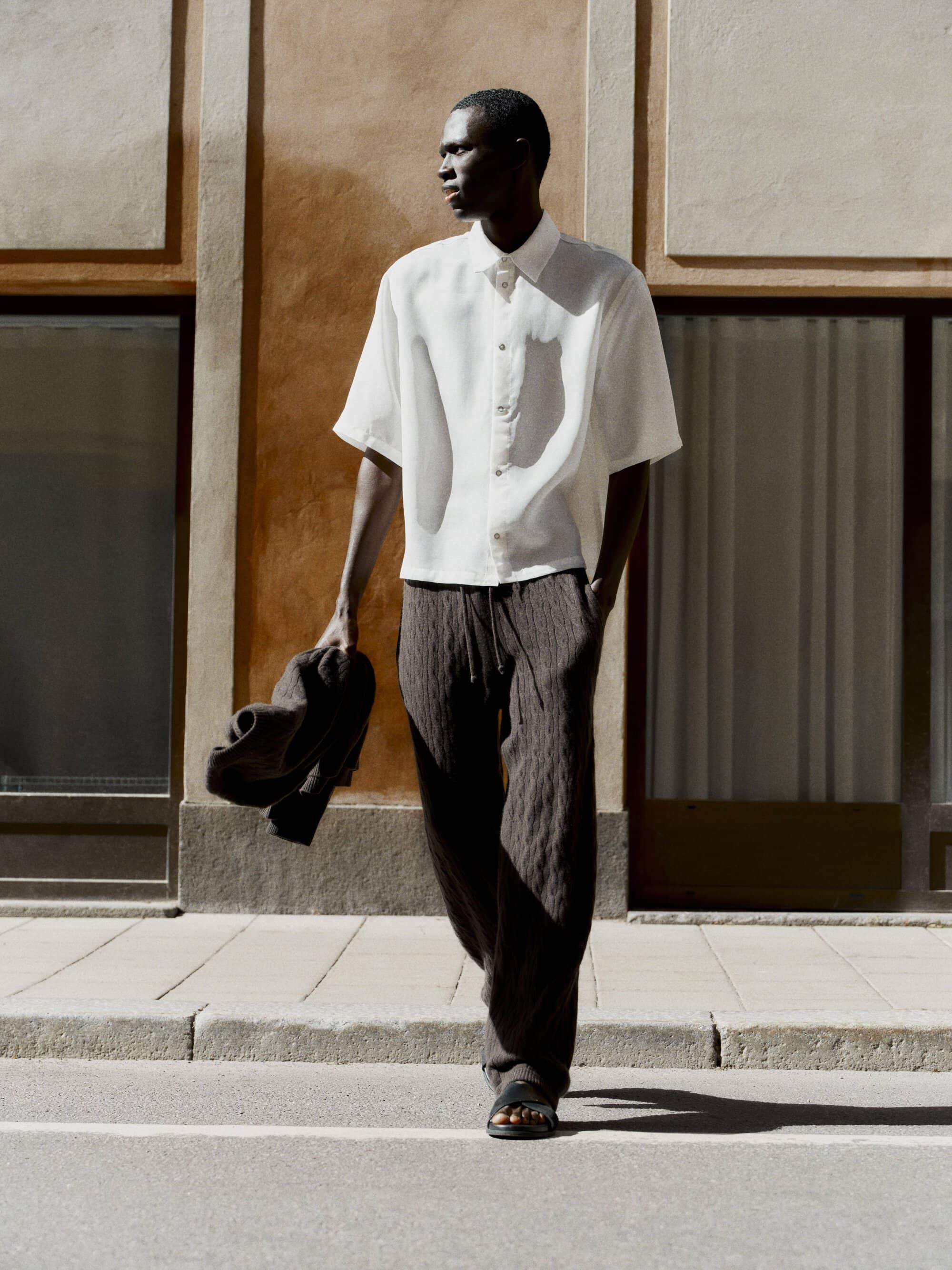 Man wearing a white short-sleeve shirt and dark trousers walking outdoors on a sunny street, carrying a jacket.