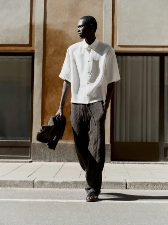 Man wearing a white short-sleeve shirt and dark trousers walking outdoors on a sunny street, carrying a jacket.