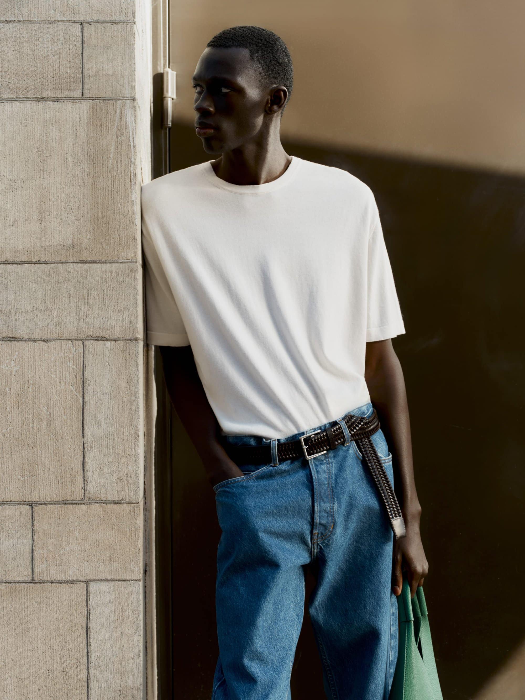 Black man wearing a white t-shirt and blue jeans leaning against a stone wall for Soft Goat fall campaign