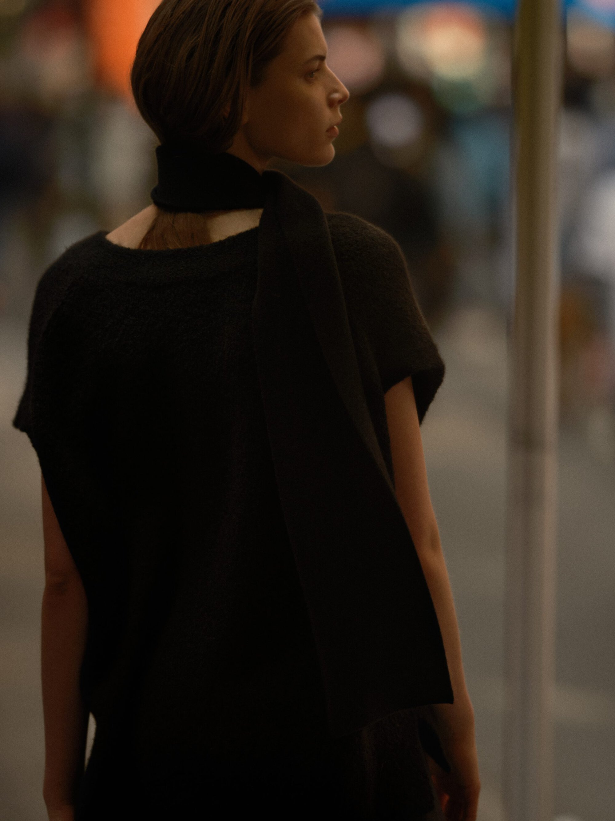 Model seen from behind wearing a black sweater with a matching scarf on a city street at dusk