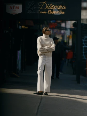 Woman in cream turtleneck and white trousers standing on a New York City sidewalk beneath a storefront sign.