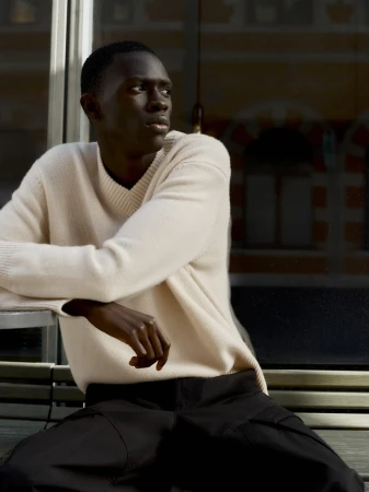 Black man wearing a cream knit sweater and dark trousers seated outdoors against an urban building background.
