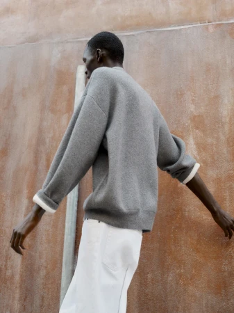 Man wearing a grey knit sweater and white trousers standing against a textured wall.