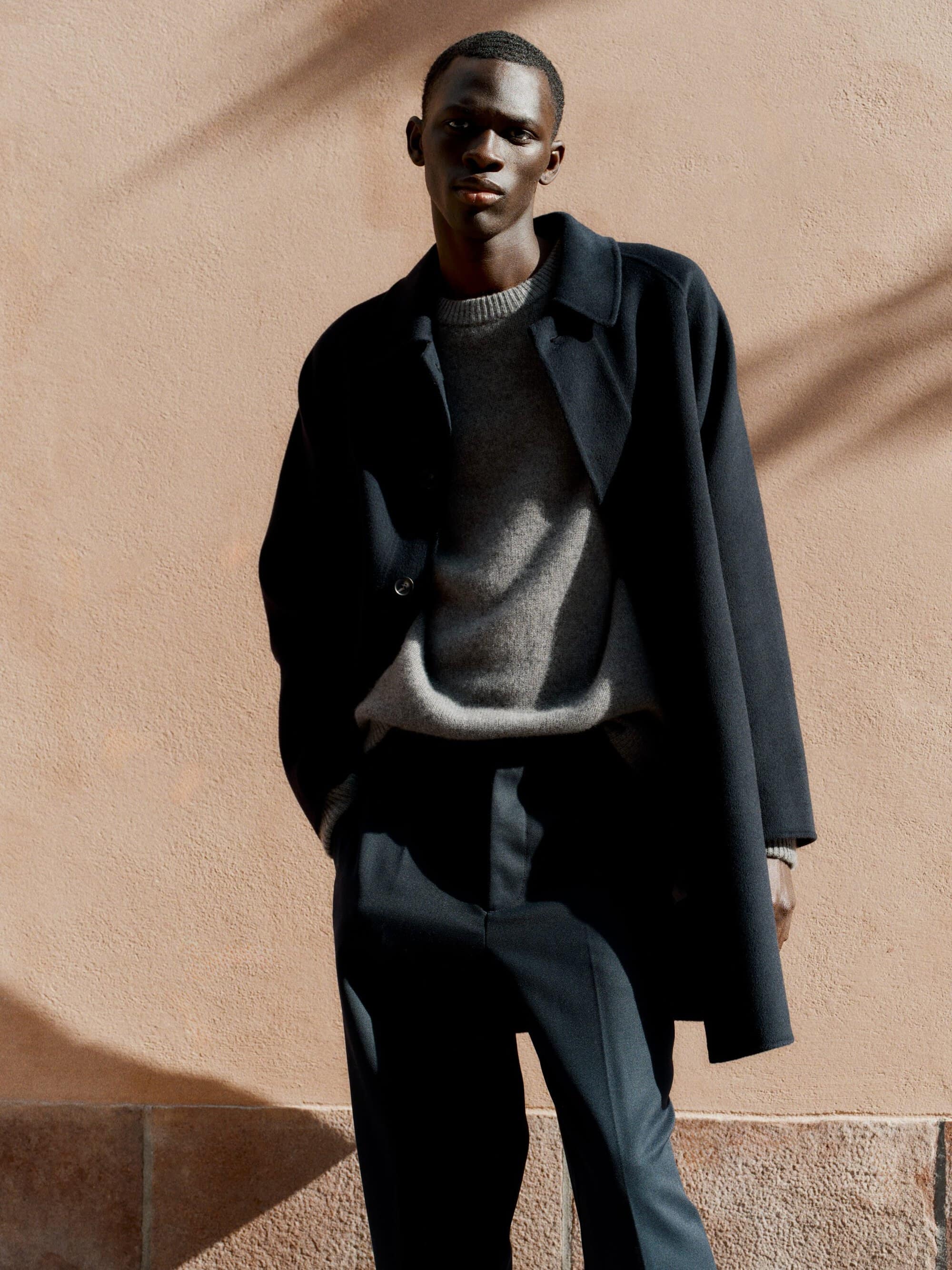 Black man wearing a dark coat and grey sweater against a textured warm background for Soft Goat Fall 2024 ad campaign