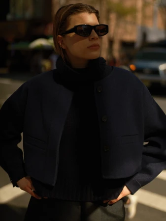 Woman wearing dark sunglasses and a dark buttoned jacket standing outdoors on a city street.