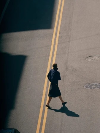 High-angle shot of a person walking across an asphalt street with double yellow lines, wearing a dark coat and skirt.