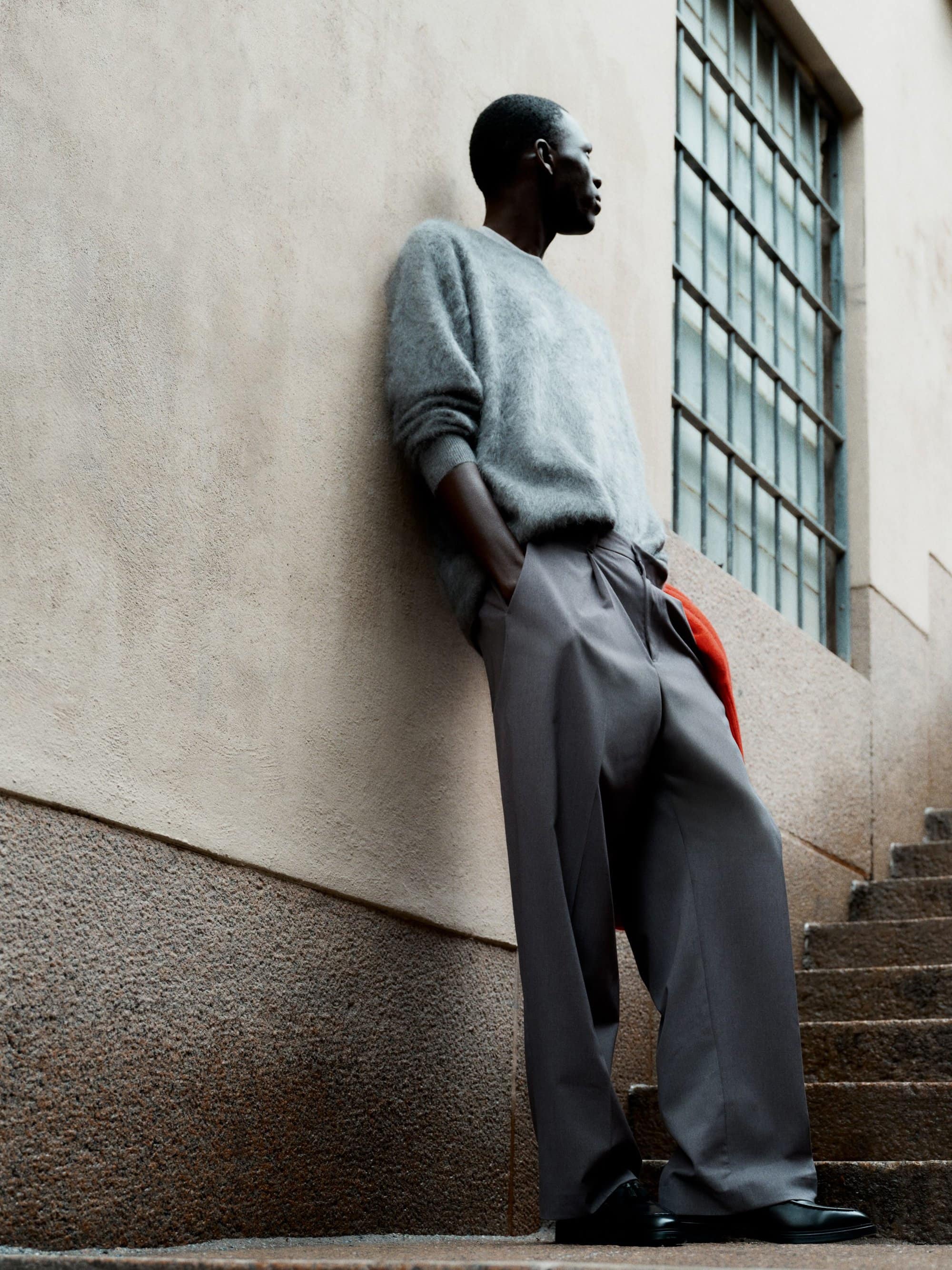 Man wearing a grey sweater and wide trousers leaning against a textured wall next to outdoor stairs