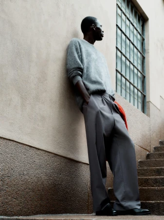 Man wearing a grey sweater and wide trousers leaning against a textured wall next to outdoor stairs