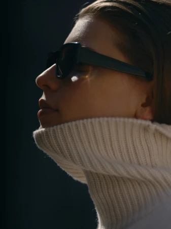 Close-up profile of a woman wearing black sunglasses and a high-collared white knit sweater in sharp side lighting.