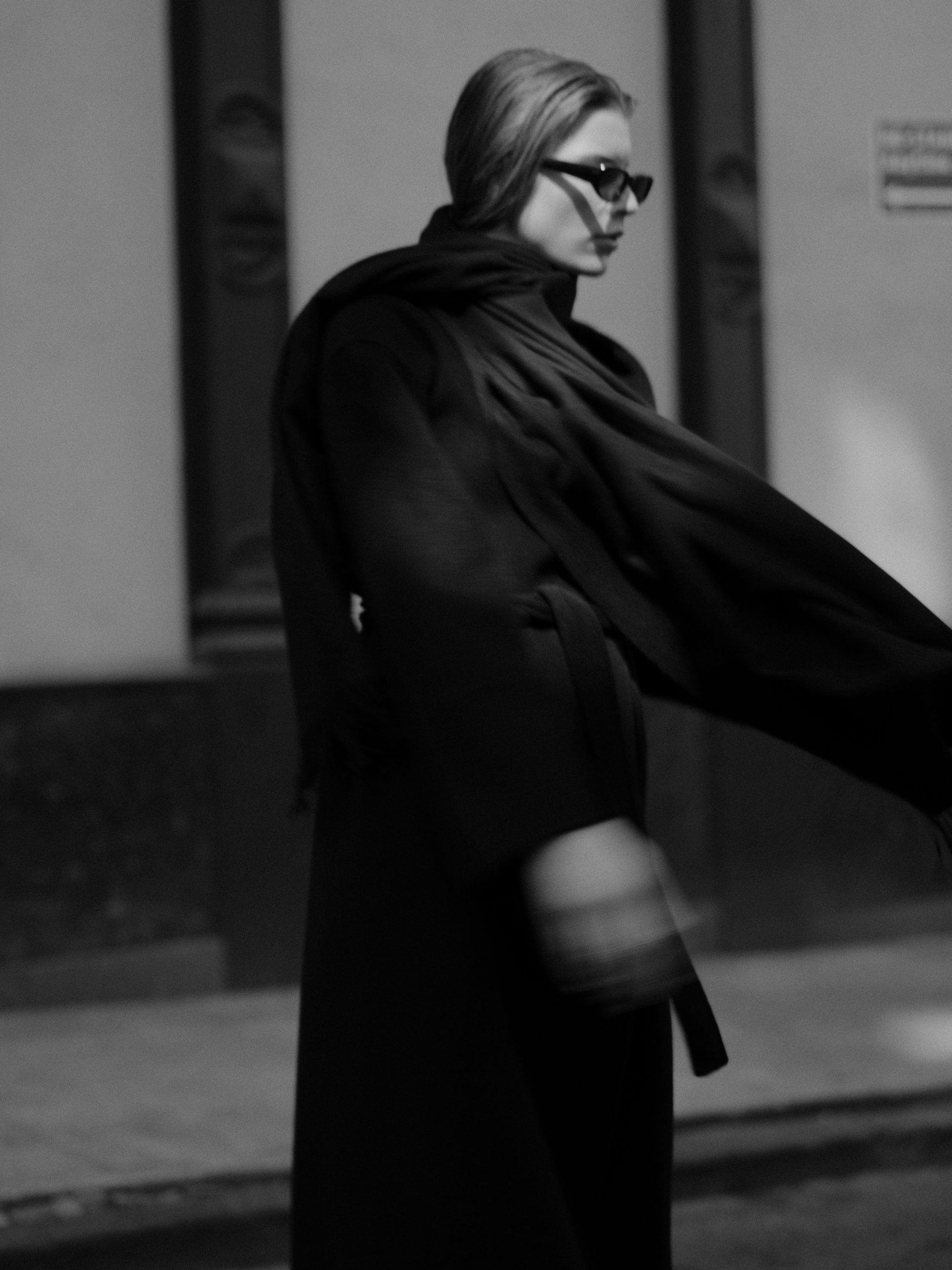 Model in dark sunglasses wearing a long black belted coat with a scarf against an outdoor urban background