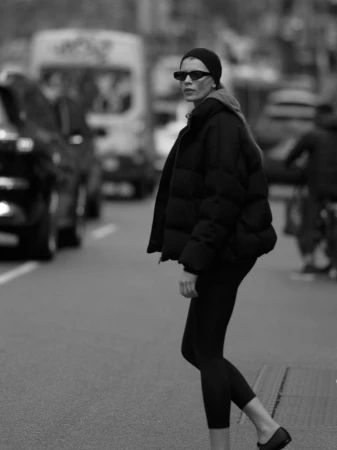 Model in black puffer jacket and sunglasses walking down a city street with out-of-focus traffic in the background.