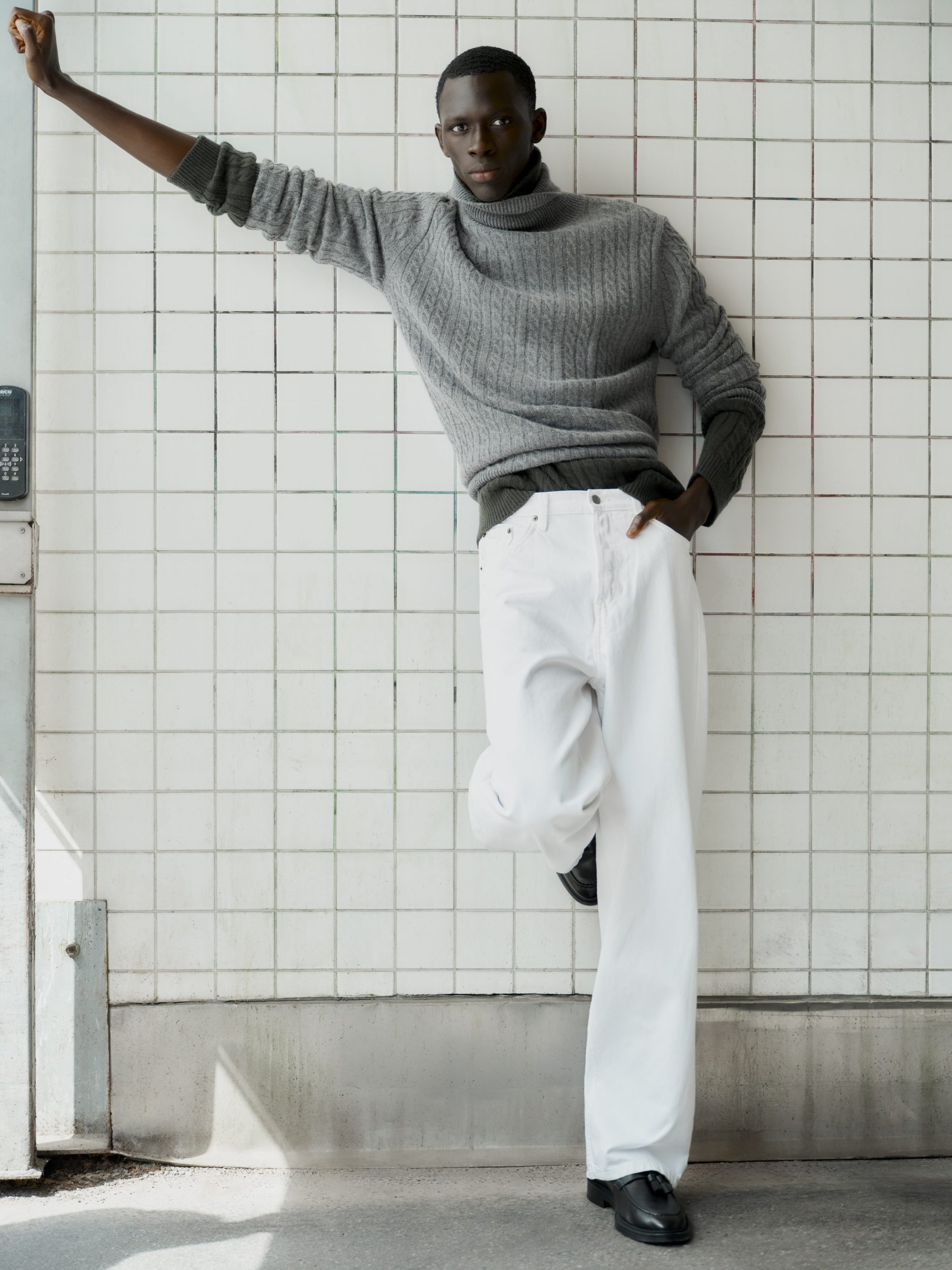 Man wearing a grey turtleneck sweater and white trousers leaning against a white tiled wall for Soft Goat Fall 2024 ad campaign