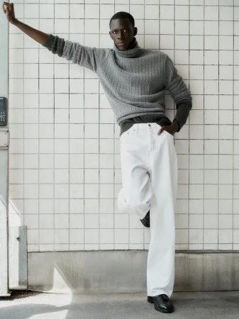 Man wearing a grey turtleneck sweater and white trousers leaning against a white tiled wall for Soft Goat Fall 2024 ad campaign