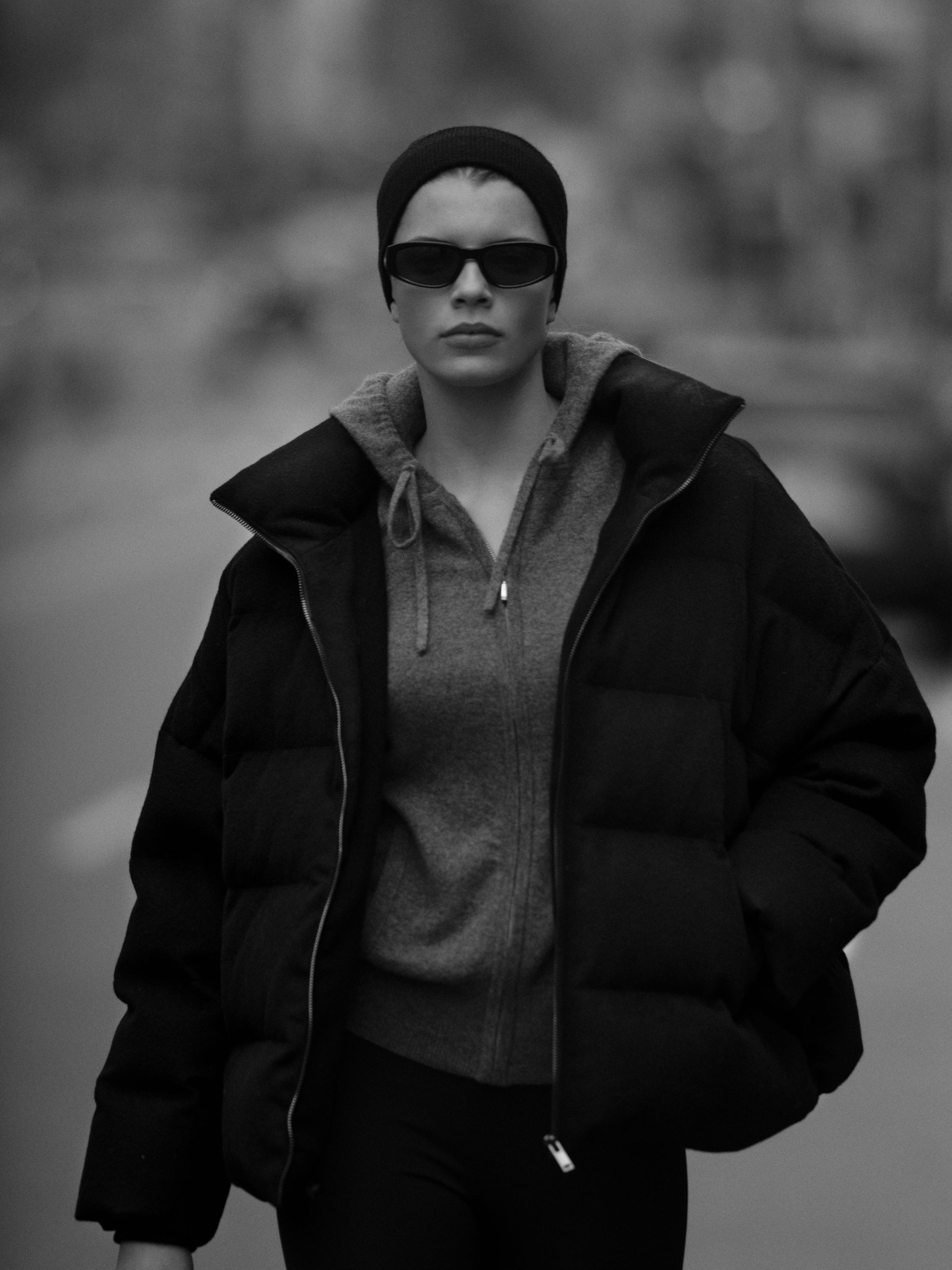 Model wearing a black puffer jacket, hoodie, beanie, and sunglasses outdoors on a city street.