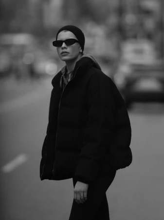 Model wearing dark sunglasses and a black puffer jacket outdoors on a city street.