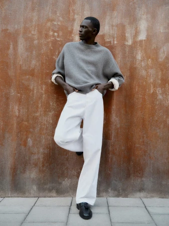 Man wearing a grey knit sweater and white trousers leaning against a textured brown wall in New York City