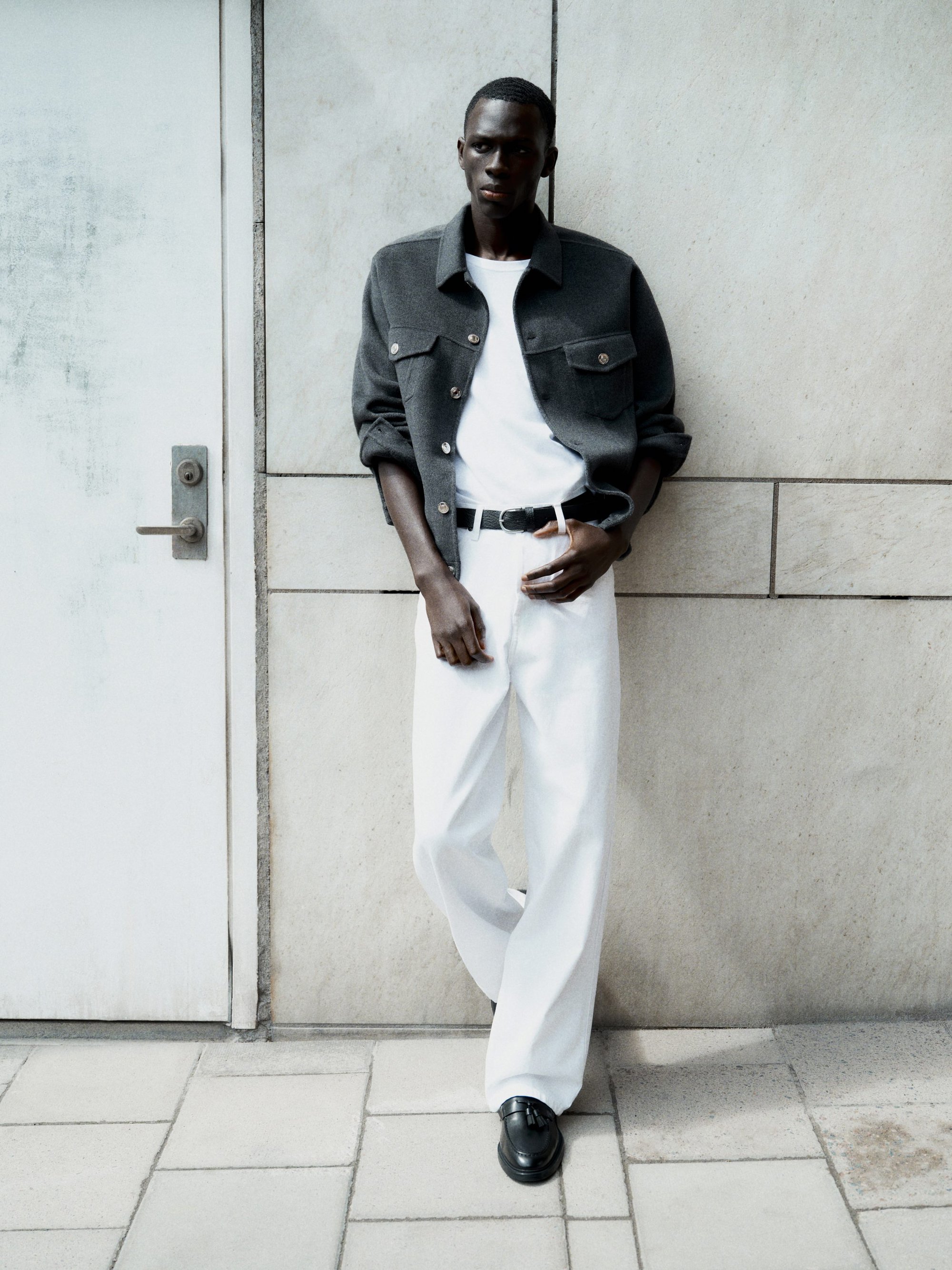 Black man leaning against a stone wall wearing a grey jacket, white t-shirt, white trousers, and black loafers.