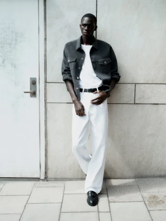 Black man leaning against a stone wall wearing a grey jacket, white t-shirt, white trousers, and black loafers.