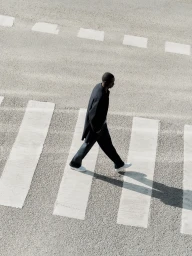 High-angle view of a man walking across a striped pedestrian crosswalk wearing a dark coat and light sneakers.