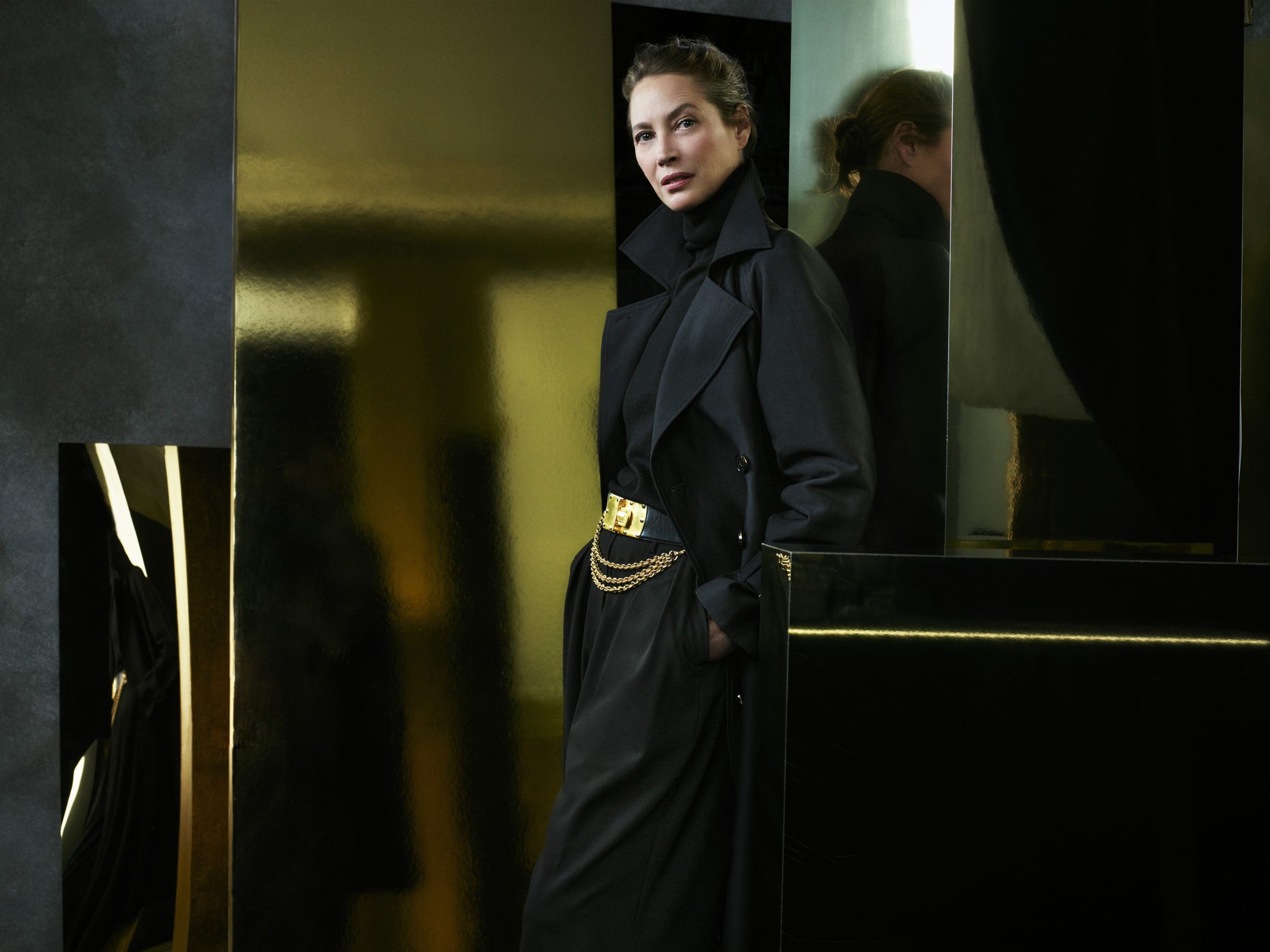 Model Christy Turlington wearing a black trench coat and chain belt against a gold and black set for Donna Karan