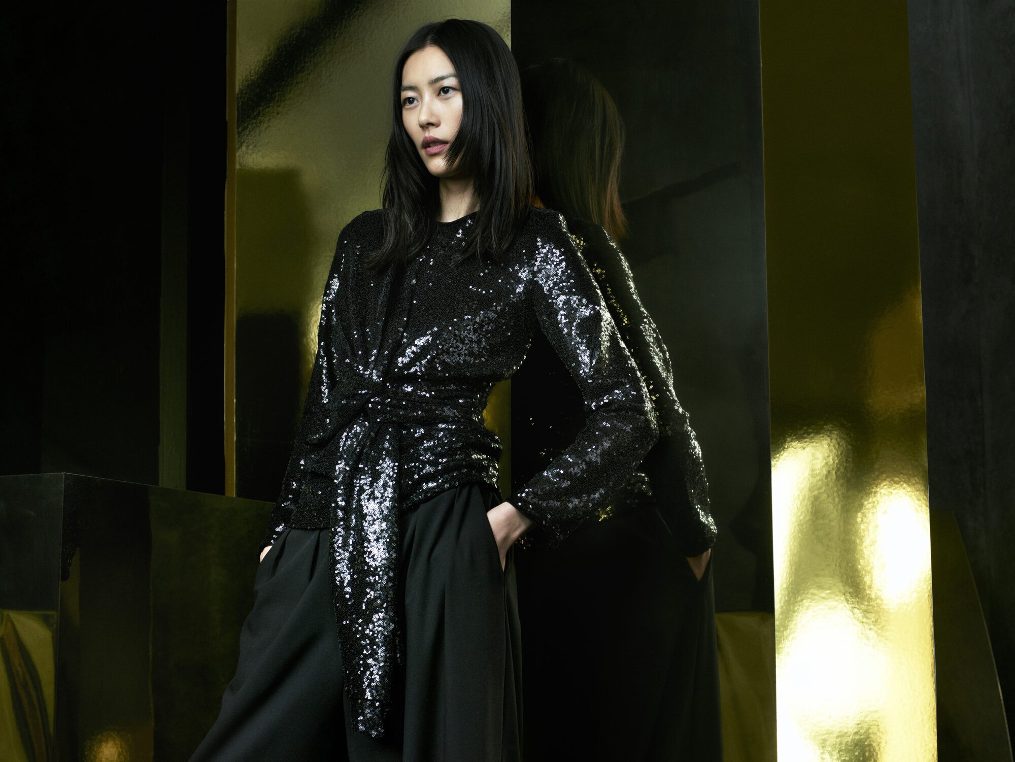 Model Liu Wen wearing a black sequin top and trousers against a dark backdrop with reflective golden panels.