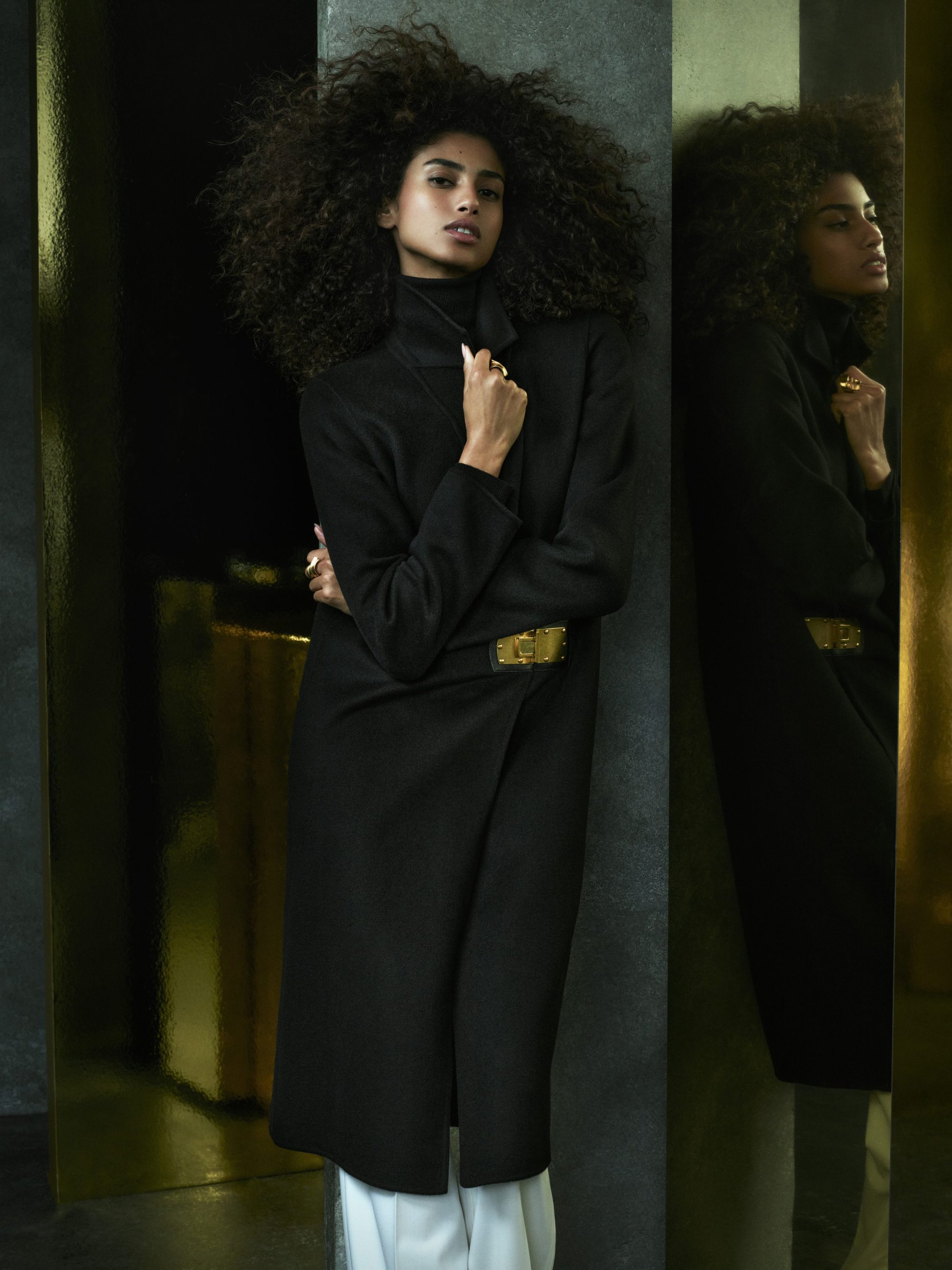 Model with voluminous curly hair wearing a black coat standing next to a reflective gold pillar for Donna Karan Fall 2024
