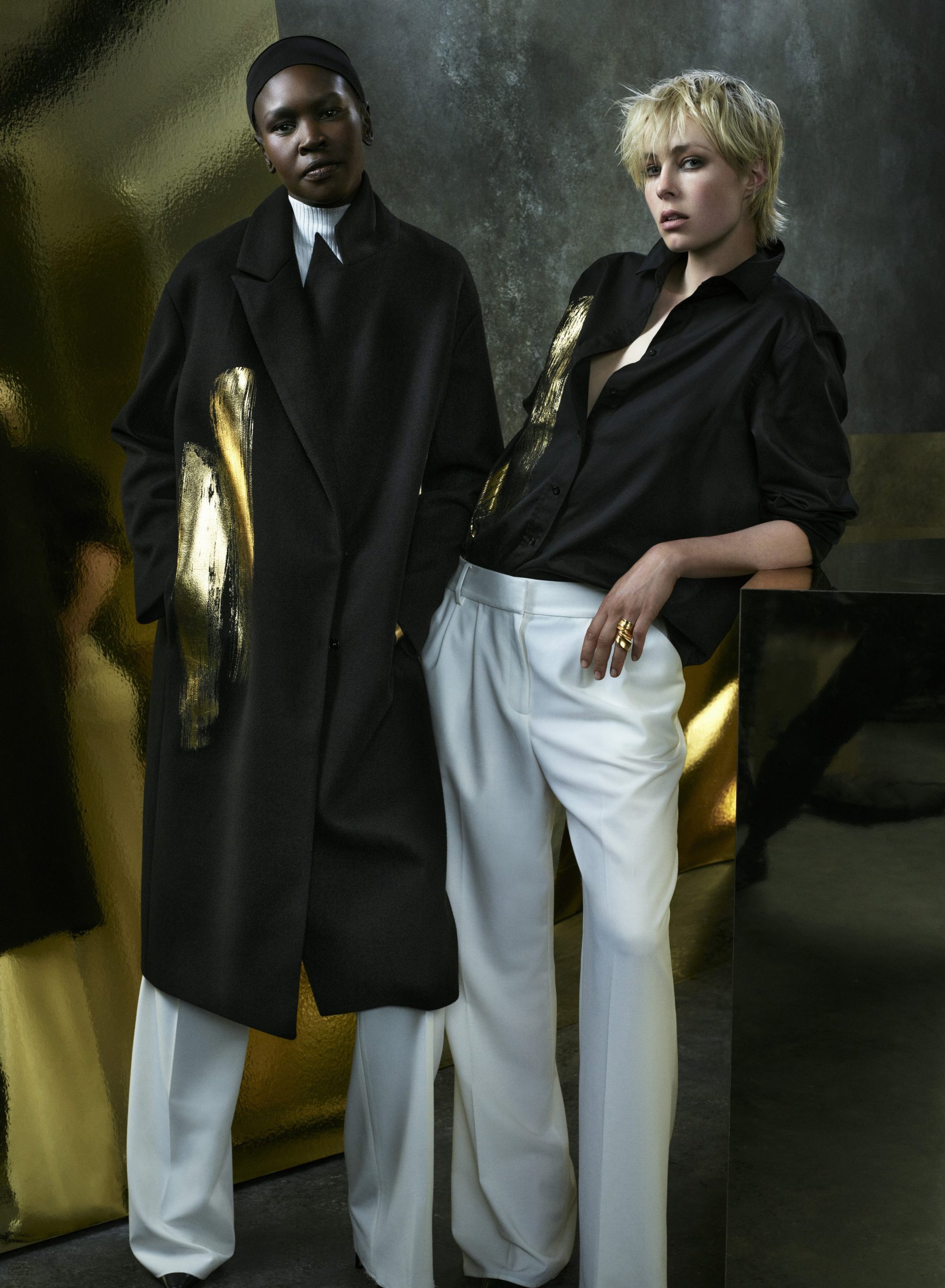 Two models posing in a dark coat and black shirt with white trousers against a textured background