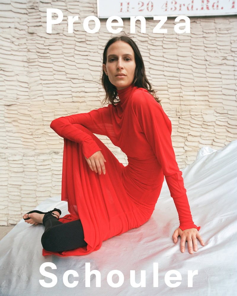 Model seated on a white draped surface wearing a long-sleeved red dress and dark shoes against a textured brick wall.