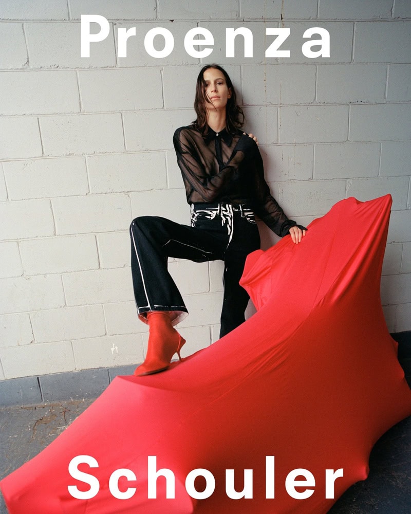 Model leaning against a white brick wall next to a draped red object wearing a sheer black blouse and dark trousers