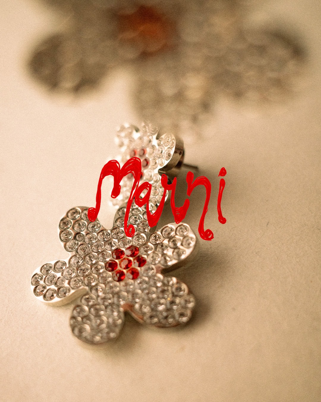 Close-up of a rhinestone flower earring with red center stones, featuring the red Marni brand logo script across the frame.