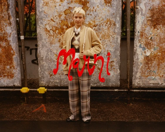 Model standing in front of weathered posters wearing a tan bomber jacket, argyle sweater vest, and plaid trousers.