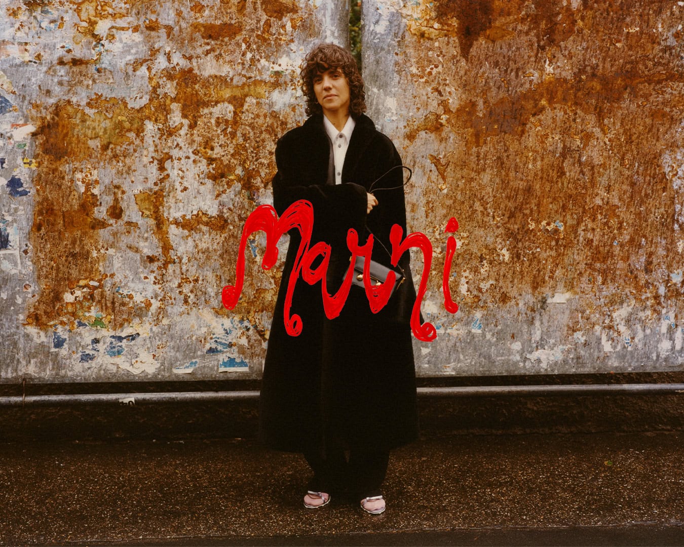 Model wearing a black coat standing against a weathered rusted metal wall with overlaid red handwritten Marni text