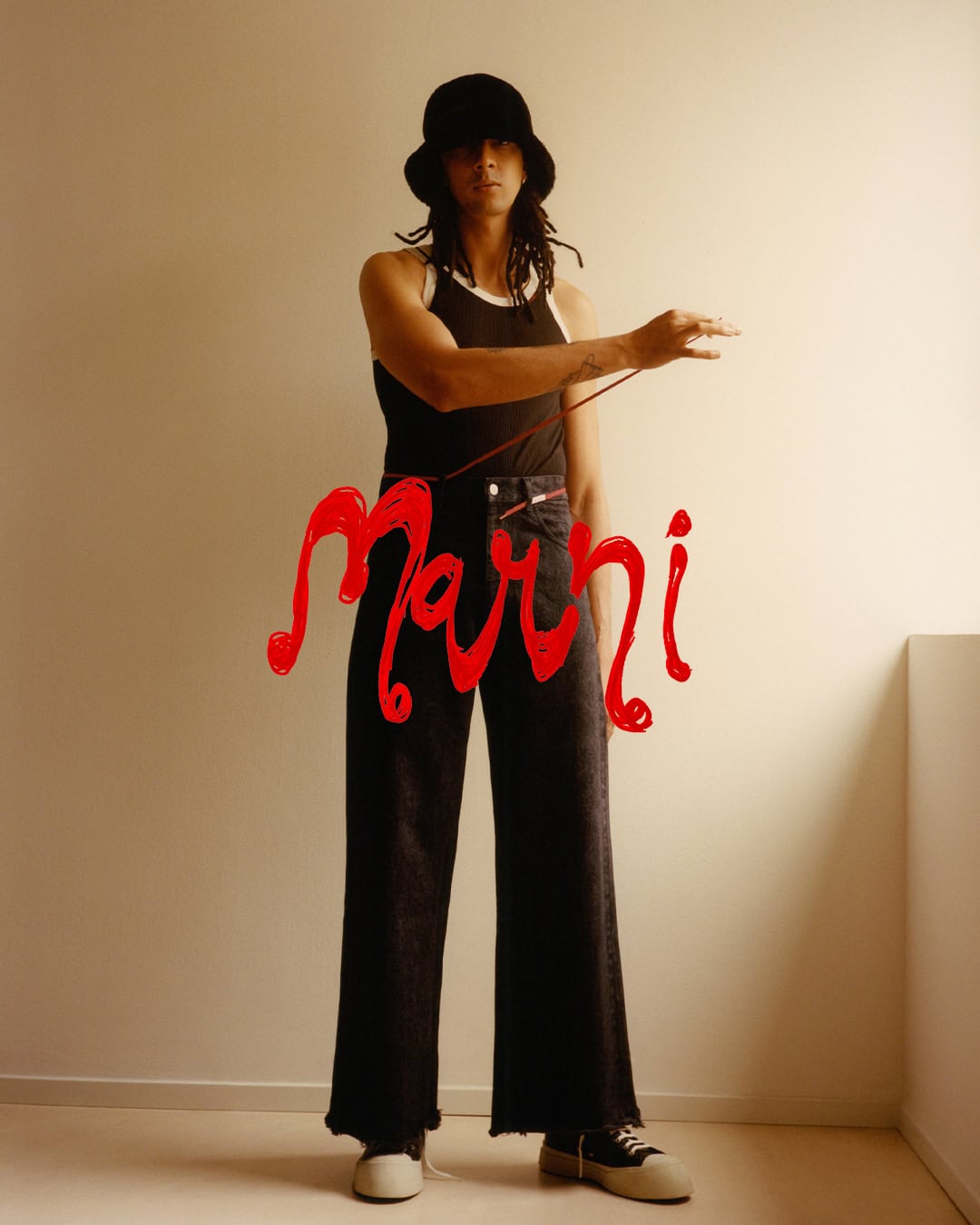 Model wearing a black bucket hat, dark tank top, and wide-leg trousers standing against a warm neutral wall in a Marni campaign image