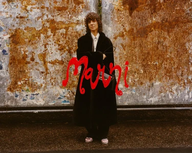 Model wearing a black coat standing against a weathered rusted metal wall with overlaid red handwritten Marni text