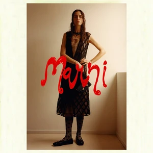 Model wearing a sheer black patterned dress and dark socks posing indoors against a plain wall for Marni