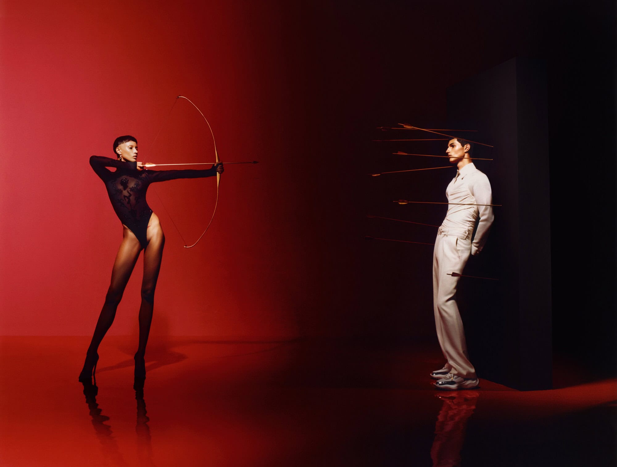 Model in black bodysuit holding a bow and arrow aiming at a man leaning against a black wall pierced by arrows