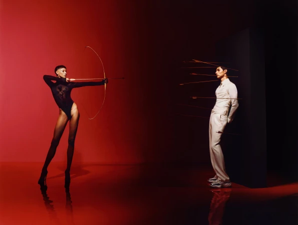 Model in black bodysuit holding a bow and arrow aiming at a man leaning against a black wall pierced by arrows