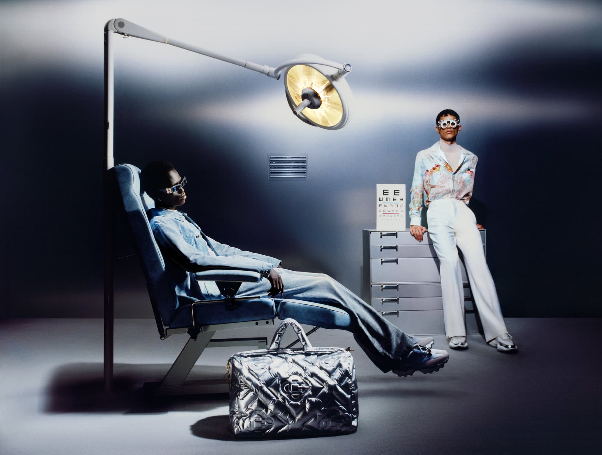 Two models in an optometry-themed studio set with a large surgical lamp, medical chair, and silver quilted bag by Casablanca.